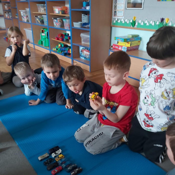 Chłopiec w czerwonej bluzeczce trzyma samochodzik, inne dzieci patrza.