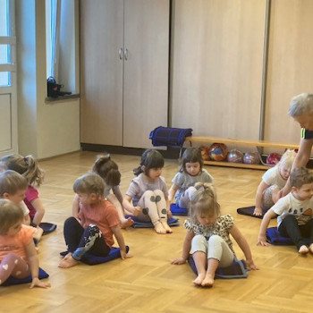 Dzieci siedzą na podłodze z wyciągniętymi nogami, pani poprawia postawę chłopca.