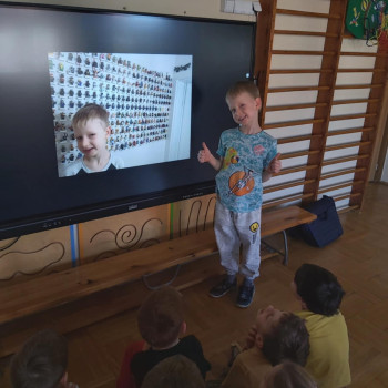 Chłopiec stoi przed tablicą multimedialną. Na tablicy jest twarz chłopca i figurki lego na ścianie. Dzieci siedzą na podłodze, patrzą i słuchają.