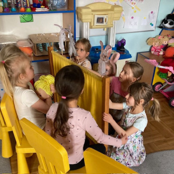 Dzieci siedzą na krzesełkach i patrza na teatrzyk. Za zasłonką dzieci pokazują kukiełki-zajączki.