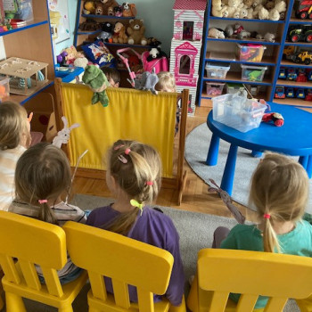Dzieci siedzą na krzesełkach i patrza na teatrzyk. Na zasłonce znajduje sie pojedyncza kukiełka, obok niebieski stolik z pudełkiem i kukiełką. W głębi regały z zabawkami.