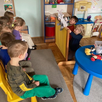 Dzieci siedzą na krzesełkach i patrza na teatrzyk. Za zasłonką dzieci pokazują kukiełki-zajączki. Obok załonki przed dziećmi znajduje się okrągły niebieski stolik, na którym leżą różne kukiełki.