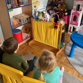 Dzieci siedzą na krzesełkach i patrza na teatrzyk. Za zasłonką dzieci pokazują kukiełki-zajączki. Obok załonki przed dziećmi znajduje się okrągły niebieski stolik, na którym leżą różne kukiełki. W głębi sali regały z zabawkami.