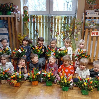 Dzieci w grupie siedzą na podłodze i pokazują swoje stroiki świąteczne.