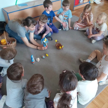 Dzieci i pani siedzą  na podłodze. Pani pokazuje wiellkanocne ozdoby - koszyczek, zajączka i pisanki.
