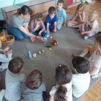 Dzieci i pani siedzą  na podłodze. Pani pokazuje wiellkanocne ozdoby - koszyczek, zajączka i pisanki.